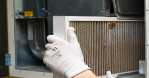 An individual in white gloves removing a dirty air filter with a white border from a piece of metal equipment.