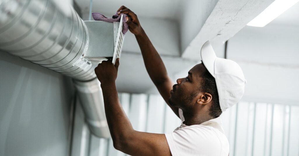 A man wearing a white T-shirt and baseball hat using a purple rag to clean a white vent connected to an air duct.