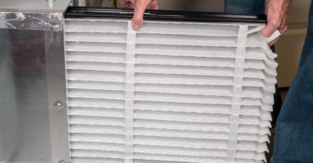 An individual in jeans installs a new white, rectangular pleated air filter into a piece of metal equipment.