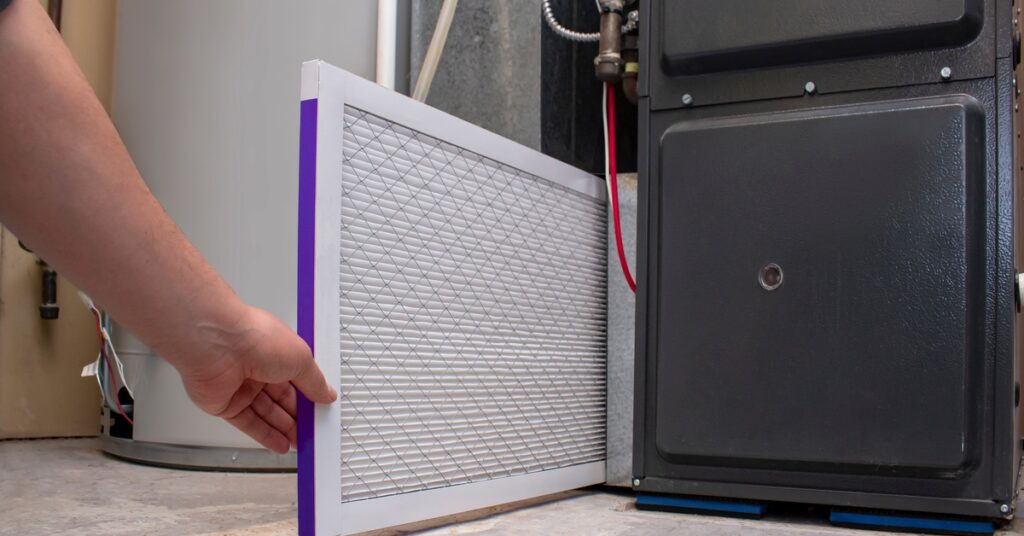 A person inserts a clean, white air filter with pleated paper and a purple border into a black furnace.