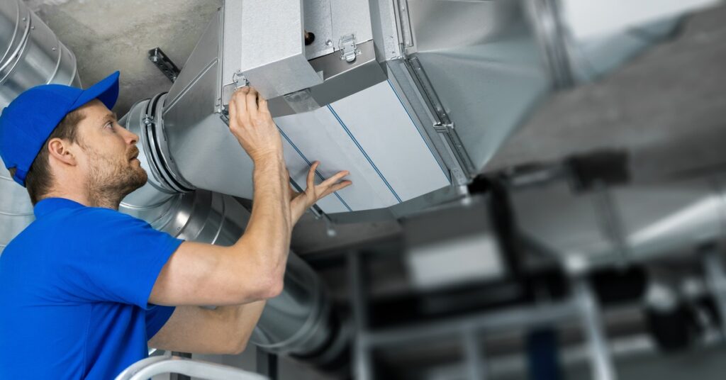 A man in a blue shirt and blue hat repairs a stainless steel ventilation system affixed to the ceiling.
