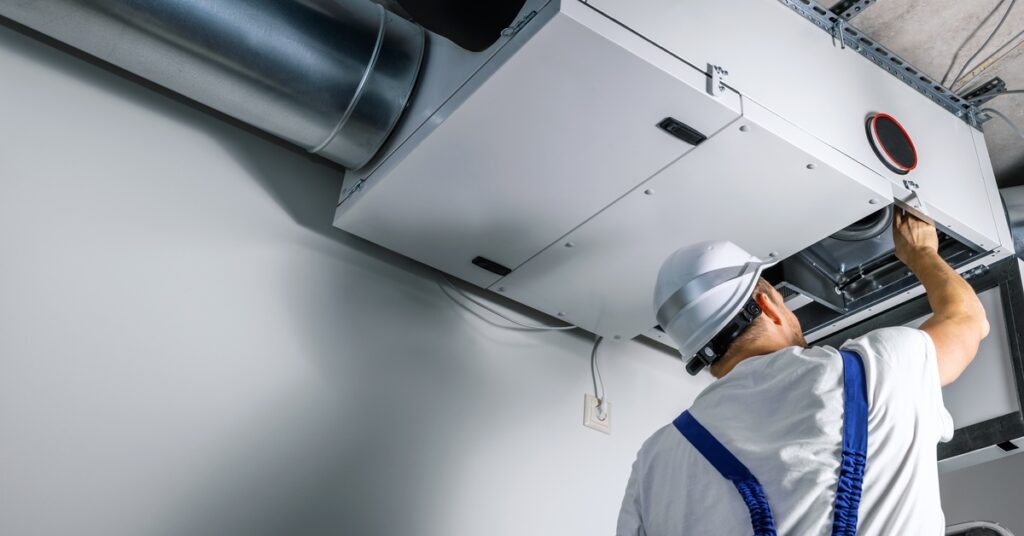 A man in a white hard hat, white T-shirt, and blue overalls services a white ventilation system affixed to the ceiling.