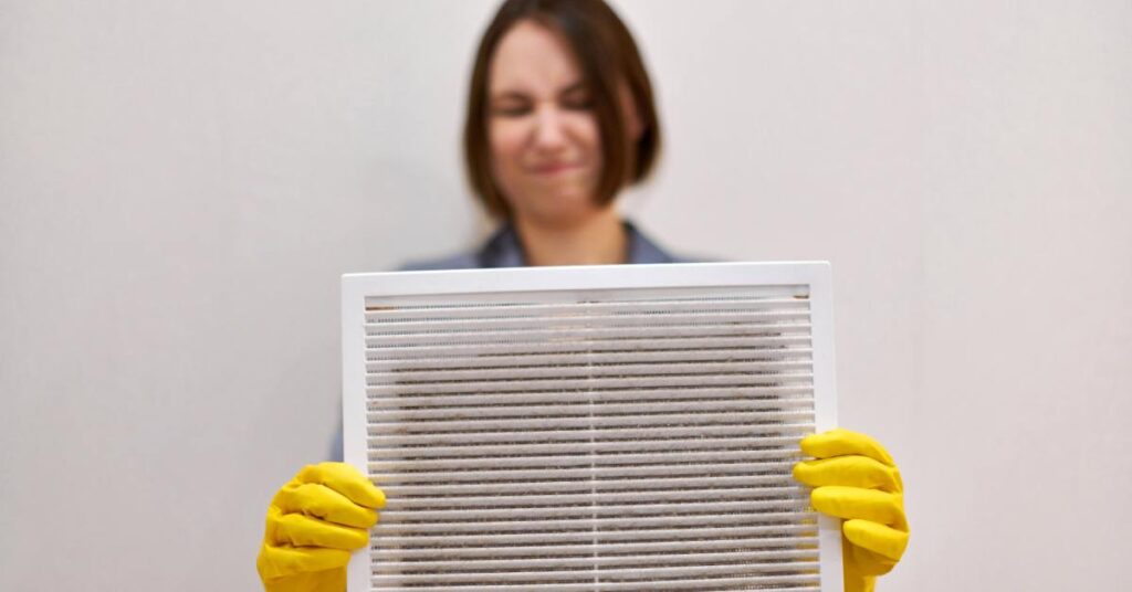 A woman with short brown hair and yellow rubber gloves holds a dirty filter with a disgusted expression.