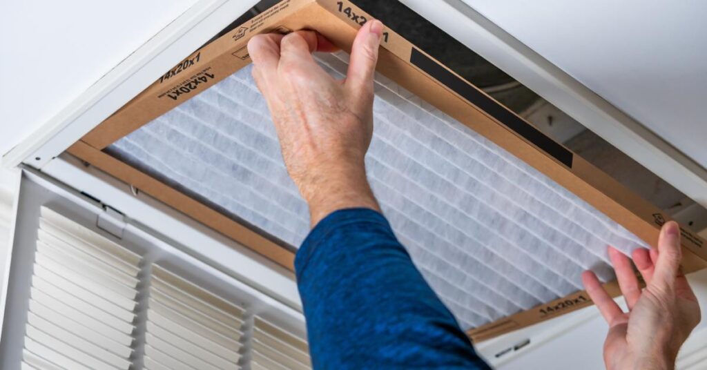 A person in a blue shirt installs a clean air filter with white pleated filter media into a ceiling vent.