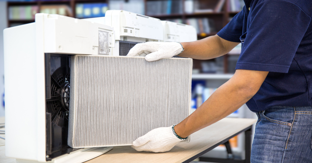 A technician in a blue shirt, jeans, and white gloves replaces a pleated filter in a large white machine.