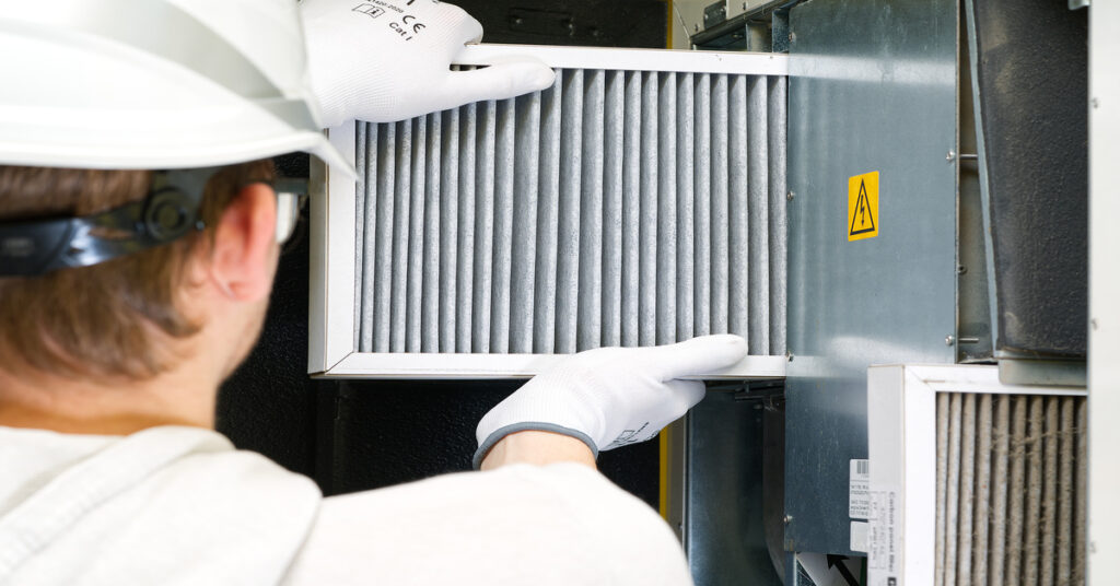 A technician wearing a hard hat, safety glasses, and gloves installs a new air filter into industrial machinery.
