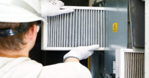 A technician wearing a hard hat, safety glasses, and gloves installs a new air filter into industrial machinery.