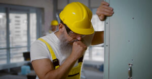 A construction worker in yellow overalls and hard hat leans on a panel while coughing in an indoor construction site.