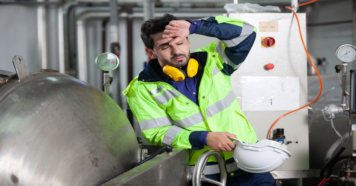 An industrial worker in a neon jacket holds his hard hat while leaning on machinery and appearing fatigued.