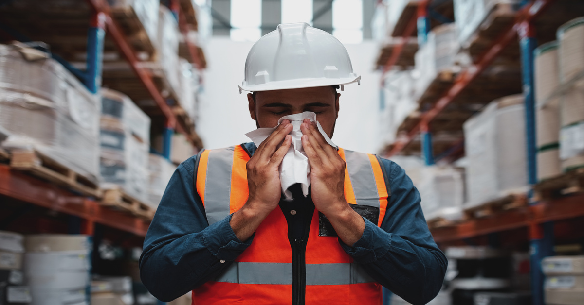 A warehouse worker in a hard hat and safety vest holds a tissue to his nose with shelves of goods in the background.