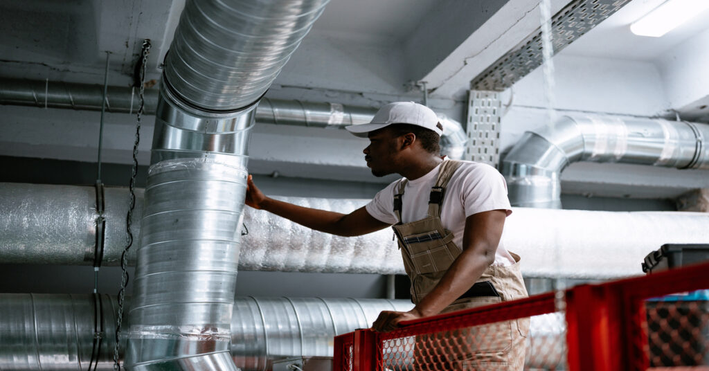 A worker in overalls inspects ventilation ducts on an elevated platform with a red safety railing in an industrial space.