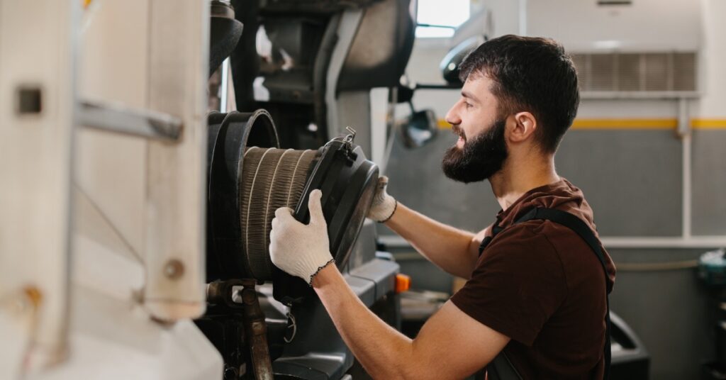 A man in a brown T-shirt, gray overalls, and white gloves replaces a large circular air filter in an industrial facility.
